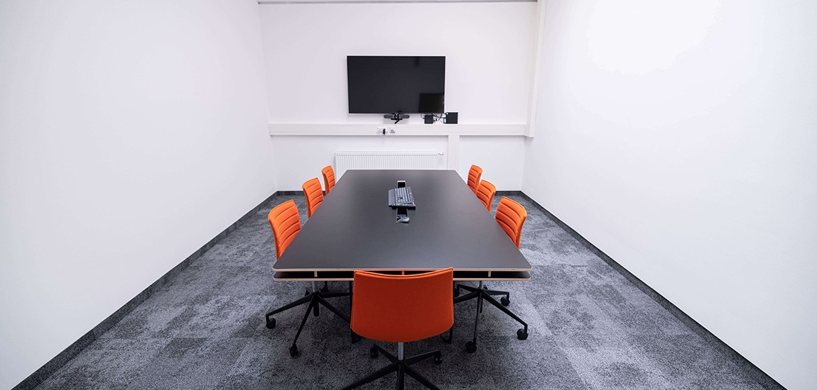 Meeting-Raum mit weißen Wänden, einem schwarzen Tisch in der Mitte um den 7 orange Sessel stehen und einem Kleinen TV an der hinteren Wand.